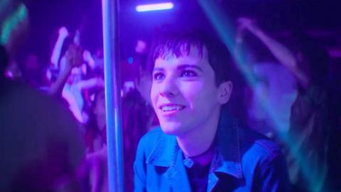 Byron stands in a club smiling. They have short brown hair and wear a blue denim shirt