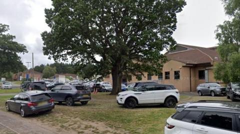 A brown brick hospital building in the background with a large tree on a grass area outside it. Cars are parked all over the grass and are not within parking spaces.