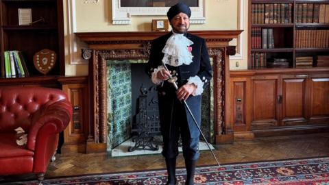 Pritpal Singh Nagi is wearing a black turban and navy blue trousers and jacket. The jacket has white frills on the chest and coming out of the sleeves, and has gold buttons. He is holding a slim sword and has long black socks on. He is standing in a regal looking room with a patterned rug of red, blue and gold. There is a red leather armchair to his left and a large fireplace with blue tiling set into a wooden wall behind him. 