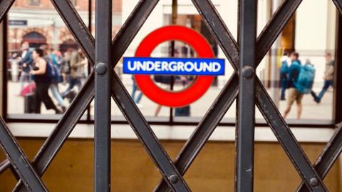 A close-up shot of a London Underground roundel, seen through the diamond patterns of a closed metal concertina gate. In the blurred background, people walk past.