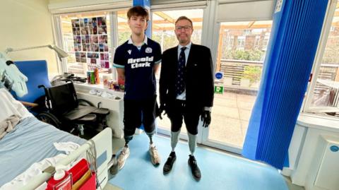 Hamish (on the left wearing a dark blue sports top and shorts with his prosthetic legs) and Craig Mackinlay (on the right wearing a short, tie and blazer with his prosthetic legs) stand in a hospital room