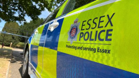 An Essex Police are parked by the side of a road. There is a cordon in place in front of the car.