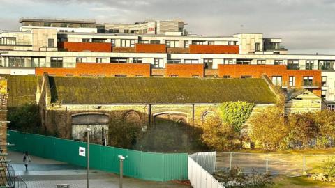 The brick railway goods shed, in the backdrop are flats. The goods shed is boarded off with green and white barriers.