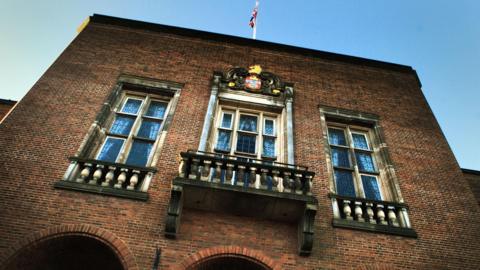 A red brick council building with three long oblong windows. A union jack is flying above the building. 