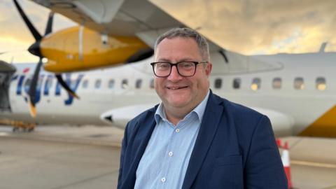 A smartly dressed man stands in front of an aircraft. He is wearing a dark blazer over a blue shirt. He is smiling at the camera. The aircraft behind him in white and yellow.