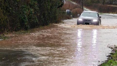 A grey Land Rover with snow on its bonnet and roof drives through floodwater which is about two feet high