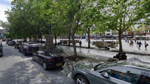 A market square lined with trees. In the foreground is a line of parked cars. People are talking, sitting and wandering around in the square.