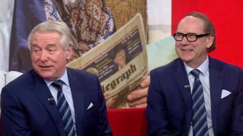 Entertainers Ian Ashpitel (left) and Jonty Stephen (right) on the BBC North West Tonight studio sofa. They are both wearing matching navy blue suits with matching ties. There is a Telegraph newspaper in the background which is part of a sketch from their show.