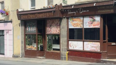 A brown shopfront of a takeaway restaurant with a sign saying China China (Town) Takeaway and Dine-In. Pictures of food on the windows including Chinese food and burgers.