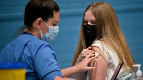 A health worker wearing a blue shirt and blue face mask holds a small cotton pad on another woman's shoulder. The other woman has long blonde hair, a black face mask and a white top.