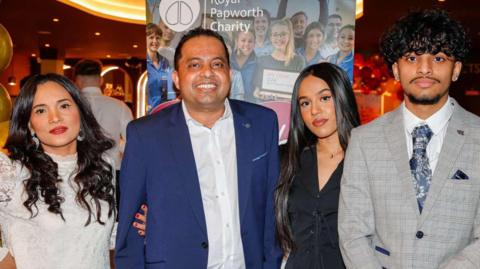 Kris Bashyal, dressed in a white shirt and blue suit, stands with his wife, wearing a white dress, daughter, wearing a black suit jacket, and son, wearing a grey suit, in front of a Royal Papworth Charity banner at the restaurant.