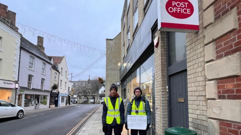 Mubashir Akhtar, right, and his son, Ibrahim Akhtar, stood outside the Co-op and post office in Malmesbury. They are wearing coats and hats, with high-vis jackets, and Mubashir holds a white piece of card with the words "Malmesbury needs post office" on it.