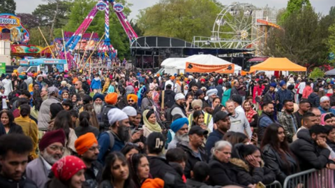 A large crowd of people looking towards something off-camera with fairground rides in the background