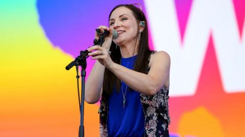 Julie Fowlis is standing on a stage at a BBC Big Weekend event. She has long dark hair, and has a pair of sunglasses on her head. She is singing into a microphone.