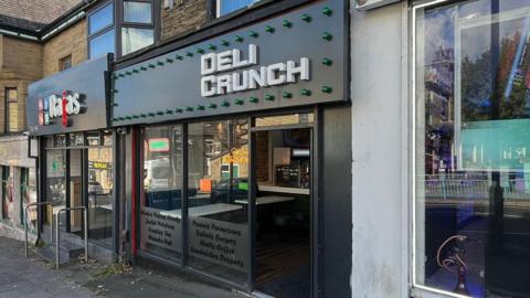 A shop called Deli Crunch sits in a row of shops. It has a glass front a black sign above saying Deli Crunch