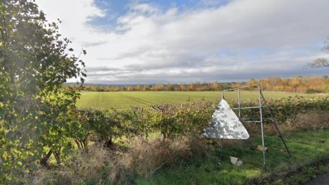 A large green field framed by a green tree and hedgerows under a cloudy sky. To the right, the back of a triangular sign resting on a trestle.