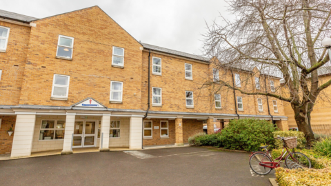 St George's Court care home is a brick building with three storeys. There is a large entrance door and a number of windows. In front is a parking area and a bicycle can be seen parked near bushes and a tree.