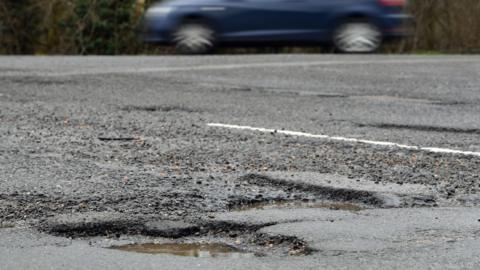 A pothole in a tarmacked road where rain has collected. Beyond it is a white line and at the top is a blurred out blue car.