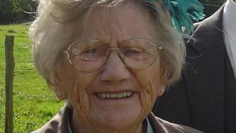 Woman with grey short hair and glasses smiles at the camera. She is wearing a green feather fascinator in her hair and stud earrings as well as oval gold-rimmed glasses. She is wearing a light brown jacket and standing next to someone else in a suit.