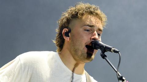 Sam Fender wearing a white tshirt singing into a black microphone with a black ear piece in his right ear.