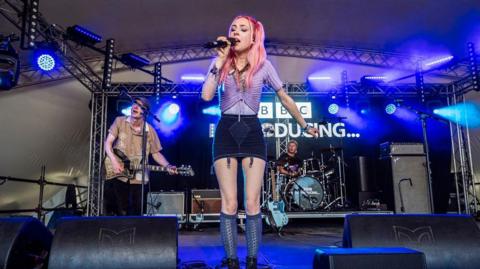 Lizzie Esau singing into a microphone on stage in front of a drummer and guitarist. She's got pink, long hair and is wearing a purple crop top and black skirt.