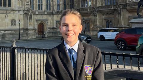 Andrii Rokotianskyi smiles as he stands outside the House of Lords. He wears a school blazer and his long, brown hair is tied back in a bun.