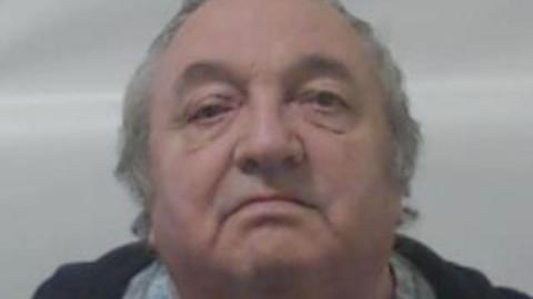 A man with thinning grey hair in a close-up police custody image. He is standing in front of a grey background. 
