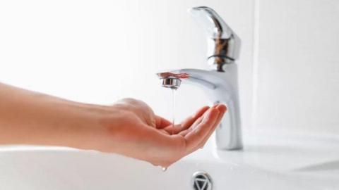 A silver tape with low water pressure. A hand is collecting the water and there are white tiles in the background.