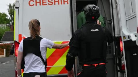 Two police officers, one in a black uniform and helmet and one in a white T-shirt and black vest, push a man in a green jumper into the back of a police van