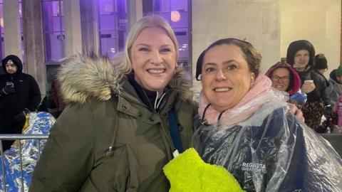 Lucy Stanbridge and Anneke Giles  who women, one is a green coat with a fluffy hood and the other is wearing a translucent poncho and holding a yellow fluffy water bottle