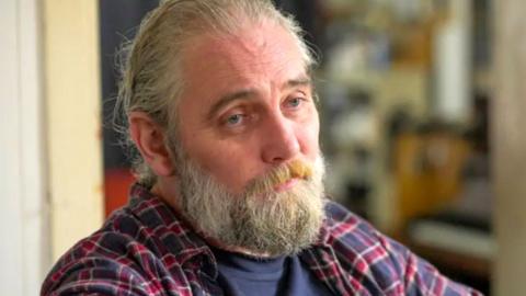 A bearded man in a check shirt is pictured sitting in is living room.