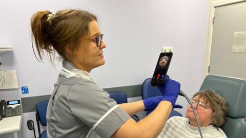 A nurse in a grey uniform holding an iPhone with a long lead attached to it and there is a woman with glasses lying in a reclining chair nearby