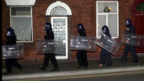 A lsingle file of police in full riot gear and holding shields walk past terraced houses on a pavement in Oldham during a second night of disturbances on May 28, 2001.