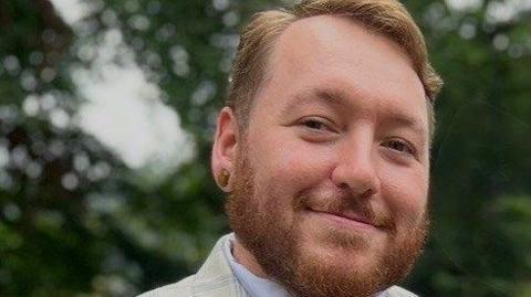 A close-cropped image of a man smiling and looking slightly to the side. He has tidy ginger hair and a beard. He has his ear pierced and is wearing a light-coloured shirt and suit jacket. the background is blurred trees or shrubbery.
