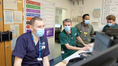 Several men in face masks sit behind computers on a desk. They wear different coloured NHS scrubs, from blue to green and behind them are posters and charts on the wall with writing on them