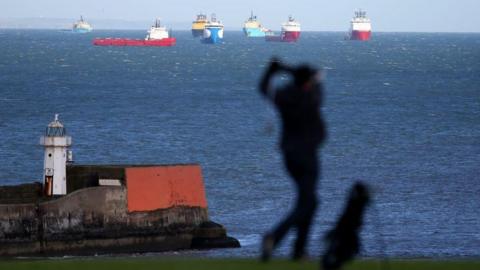 Silhouette of a golfer, with a lighthouse in the background, and offshore industry vessels in the sea.
