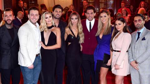 Members of The Only Way Is Essex cast on a busy red carpet at an ITV gala. They are all dressed in smart clothes, with the men in suits and women in dresses. They are all smiling at the camera.