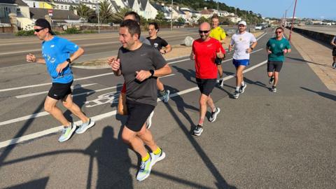 A group of eight people running along a road on a sunny day. They are all wearing t-shirts and shorts.