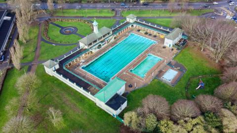 A drone image of the Peterborough Lido. The site features a large and small swimming pool with shrubs, trees and paving around the perimeter. 

