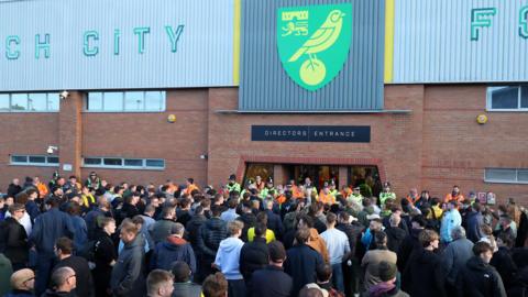 A crowd of around 50 football fans are stood outside the doors to a stadium, with a sign above describing it as the directors' entrance.