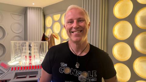 A man with short blond hair is smiling for a head and shoulders photo and is wearing a black top that says The Console Room on it. He is also wearing a circular necklace on a black thin rope. Behind him are grey walls with yellow circles on it, a hat stand with a hat and brown coat and a scarf on the other side. Next to him is a clear cube on a console with several buttons.