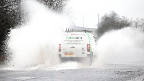 A white van drives through a large puddle in the road. Spray comes off each side of the vehicle. To the right of it the spray is vehicle high. To the left the spray is twice the height of the vehicle. There is a leafless tree to the left of the van. 