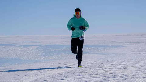 Paul Holborn running in Antarctica on the snow. He's got his colourful glasses on with a light blue hoodie and long, black trousers.