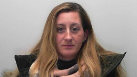 A pink-faced woman with long brown and blonde streaked hair, and a large brown coat collar, stares into the camera.