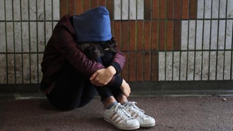 A stock shot of someone sitting on the ground in a subway. They have their arms around their knees and their head is resting on their knees. They are wearing white trainers, a blue top and trousers. Their hood is up, covering their face.