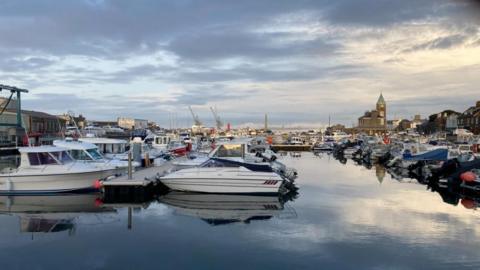 An image of white boats moored in rows St Sampson's harbour in Guernsey. The water is still and reflecting a cloudy sky, in the background is a clock tower and cranes, as well as brown building. 