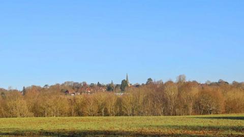 A shot of rural land showing a field, woods and a village in the distance with church spire on a sunny day.