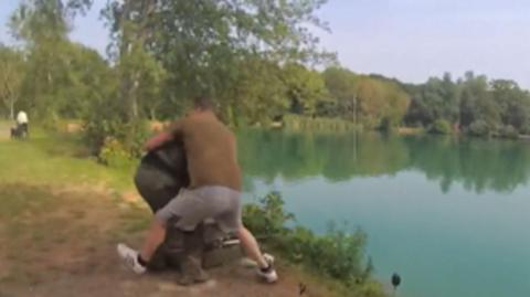 A man in grey shorts and a brown top wrestling a man wearing green trousers and a dark top, by the side of a lake. The image is blurred.