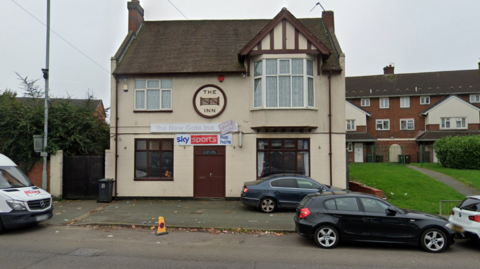 The pub is next to houses with cars parked outside the front of the premises. It is a small building and has a brown front door with a Sky Sports sign above it and the pub's name above that.