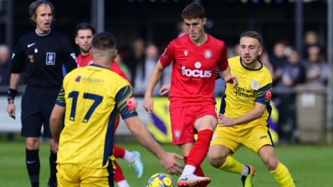 Jack Wadham passes the ball with his instep in a crowded midfield, wearing Worthing's all red home kit